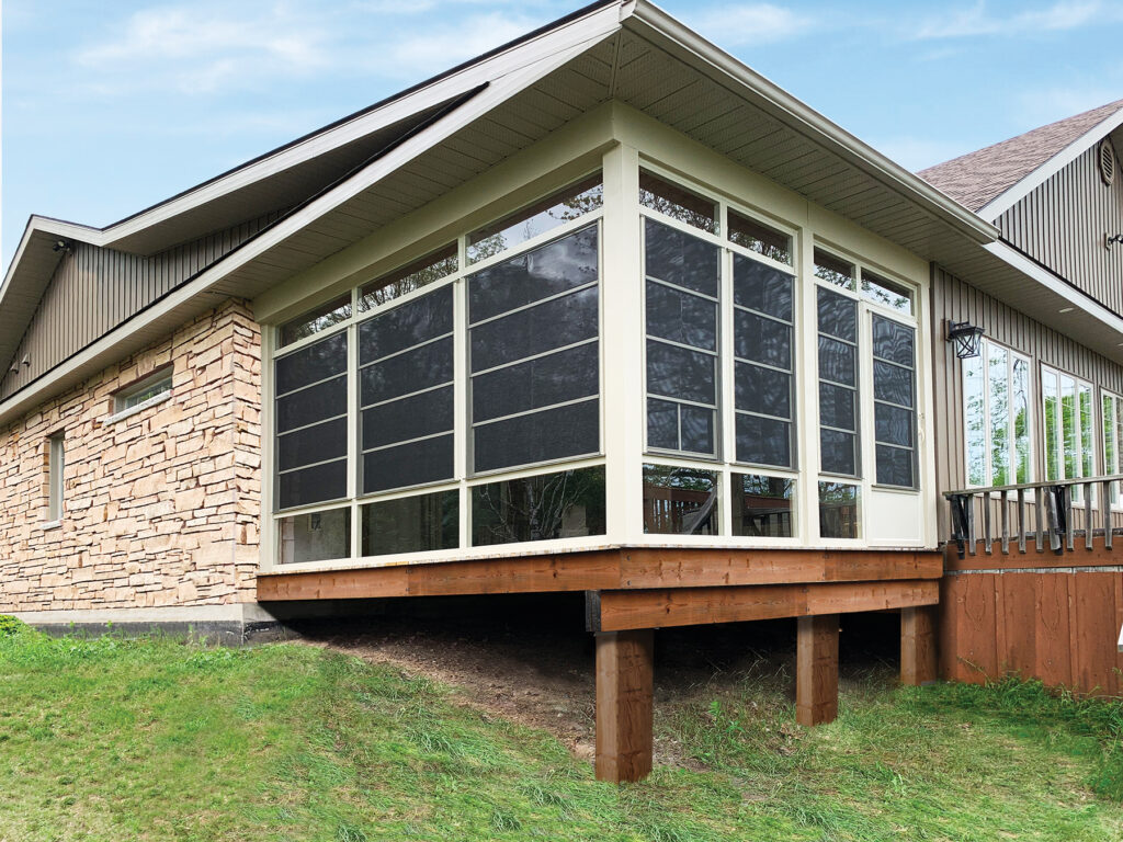 Exterior of a tan-framed sunroom built on a wooden deck featuring 4-track stacking vinyl windows partially opened for ventilation.