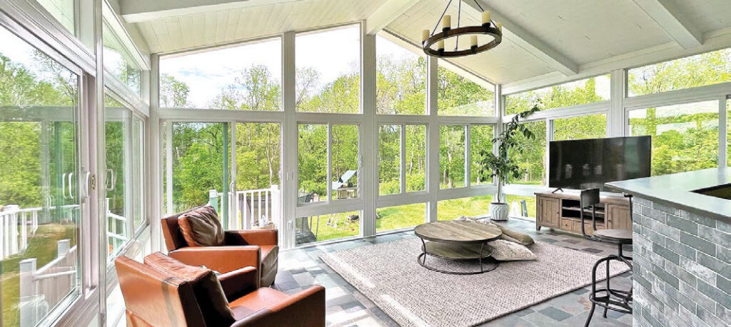 Interior of a bright 4-season sunroom featuring large windows, a vaulted white ceiling with a chandelier, leather armchairs, and a large TV.