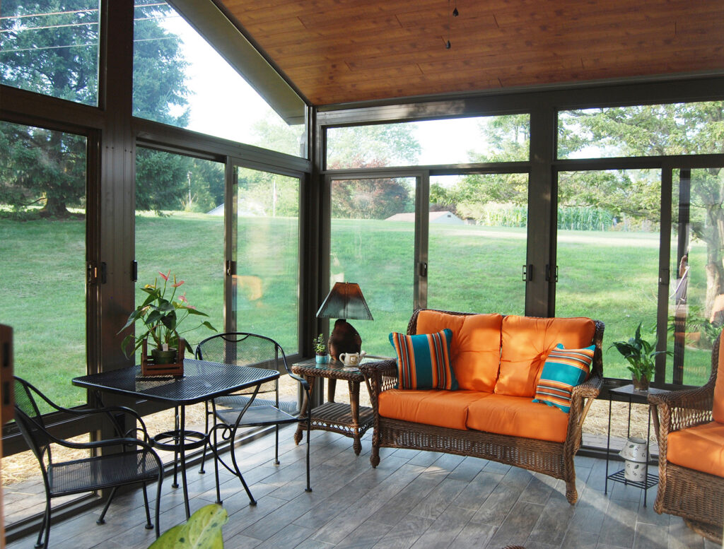 Interior view of a premium 3-season sunroom featuring a wood-finished ceiling, large glass sliding doors, and comfortable patio furniture.