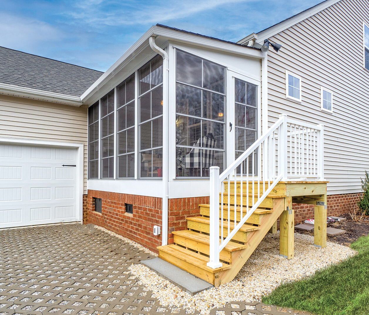 Custom Deck Builder for Homeowners - M2 Decks & Enclosures Exterior view of a white 3-season sunroom with 4-track stacking windows, a brick base, and wooden entry stairs.
