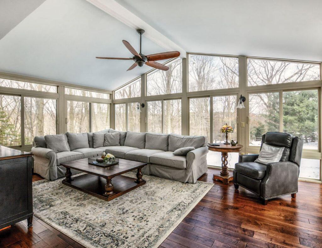 Interior view of a 4-season sunroom in winter, featuring energy-efficient glass walls, a vaulted ceiling with a fan, and a comfortable living room setup.