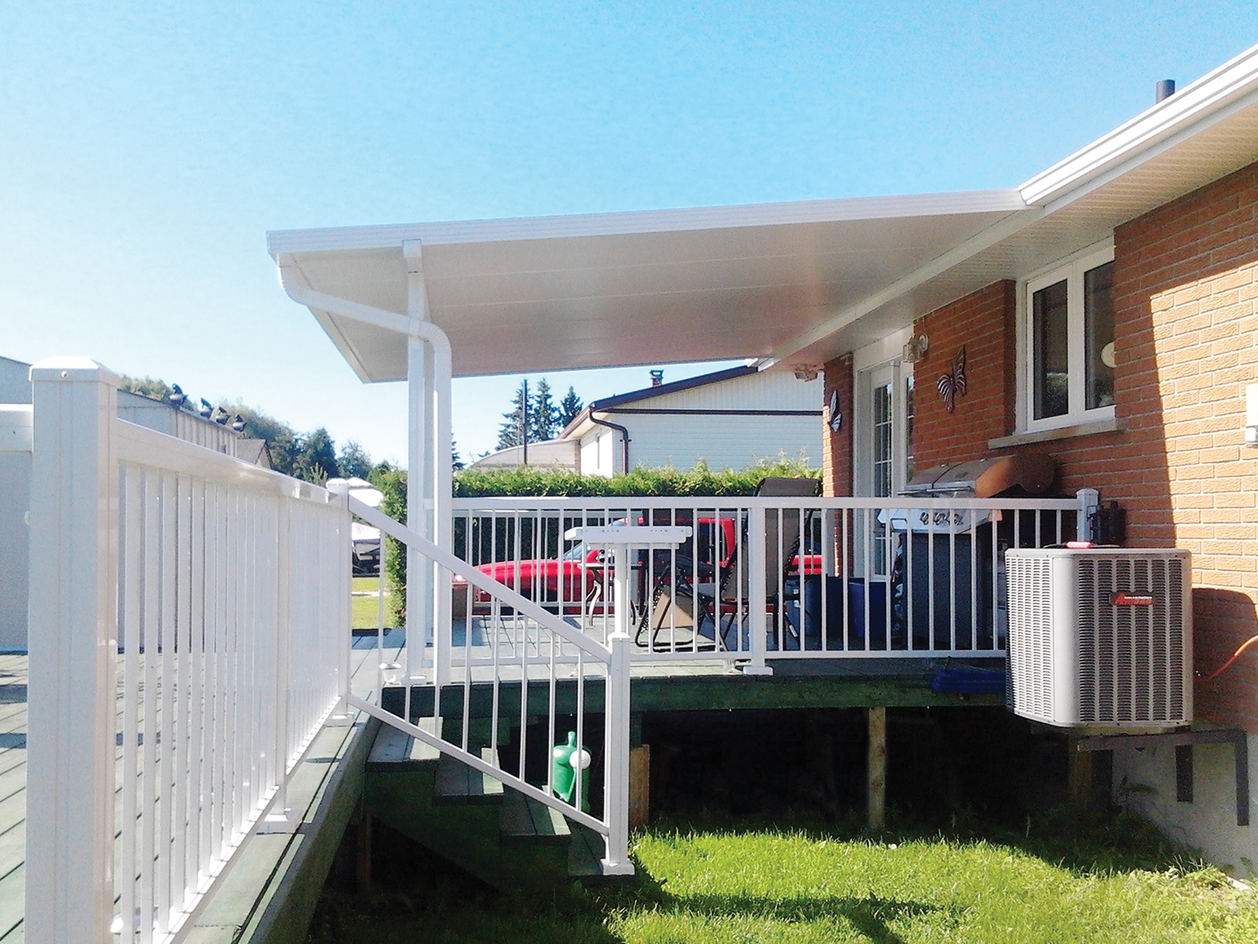 Best Deck Company - M2 Decks and Enclosures A white structural aluminum patio cover installed over a raised wood deck with white railings, featuring a grilling station and outdoor seating area on a brick home.