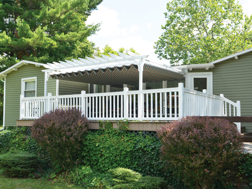 A white aluminum pergola with traditional open rafters and an integrated retractable striped fabric shade, installed on a deck with white railings.