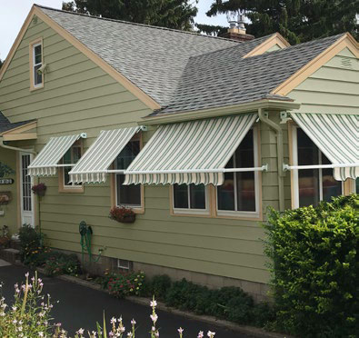 Custom Deck Builder for Homeowners - M2 Decks & Enclosures Four green and white striped retractable window awnings installed on a sage green home to block solar heat gain and protect the interior.