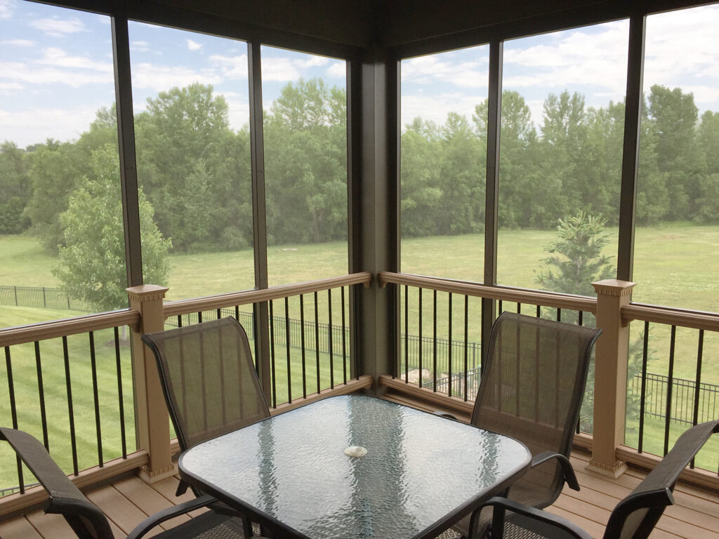 Interior of a custom aluminum screen room with a glass-top dining table, brown chairs, and a scenic view of a green Wisconsin backyard.