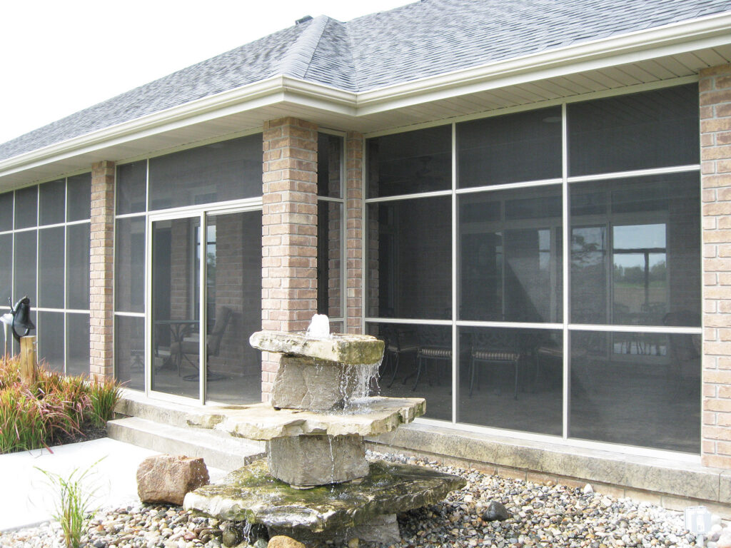 Exterior view of a large white aluminum screen room with a sliding door, integrated into a brick home with a stone water fountain in the foreground.