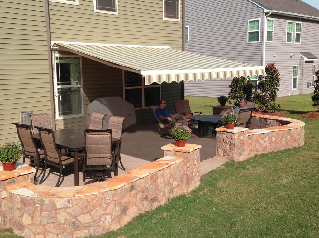 A striped tan and sage green retractable awning extended over a stone patio with a dining set and lounge chairs.