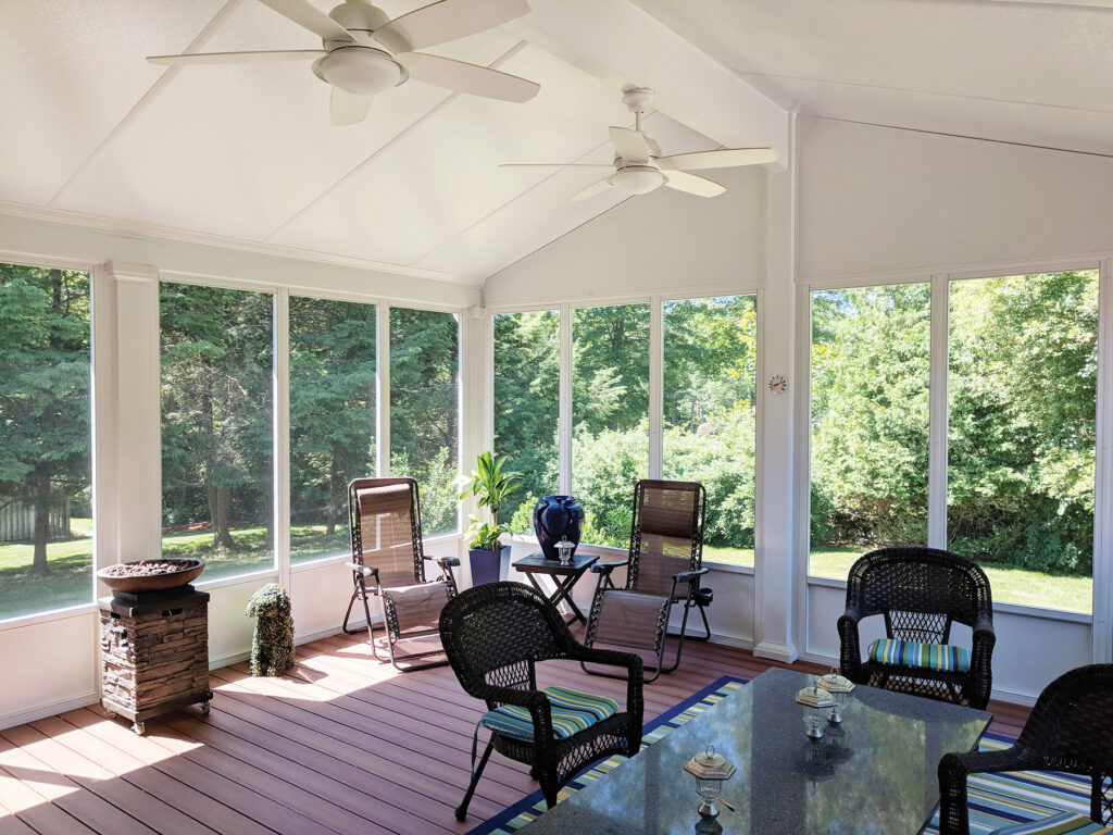Interior of a furnished white screen room with two ceiling fans, wicker chairs, a fire pit, and a dining table overlooking a wooded yard.