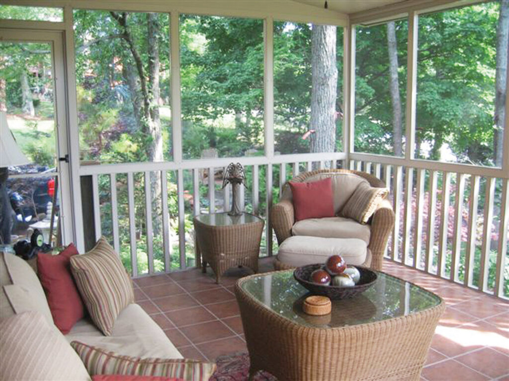Interior of a cozy, tan-framed screen room with wicker furniture, terracotta tile flooring, and high-visibility mesh overlooking a lush, wooded backyard.
