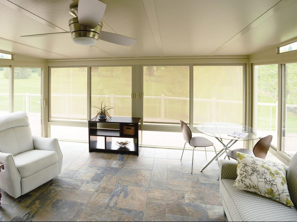 Custom Deck Builder for Homeowners - M2 Decks & Enclosures Interior view of a sunroom with slate tile floors and a ceiling fan, showing vertical solar shades lowered to reduce glare while maintaining a view of the backyard.