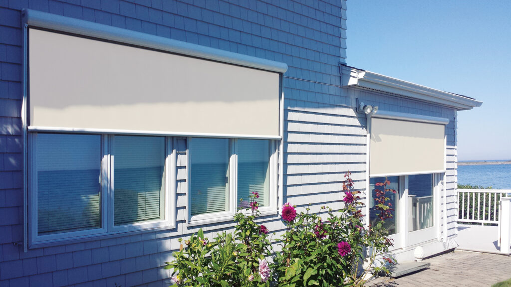 Two tan motorized solar shades installed on the exterior of a light blue shingled lake house to protect large windows from water glare.