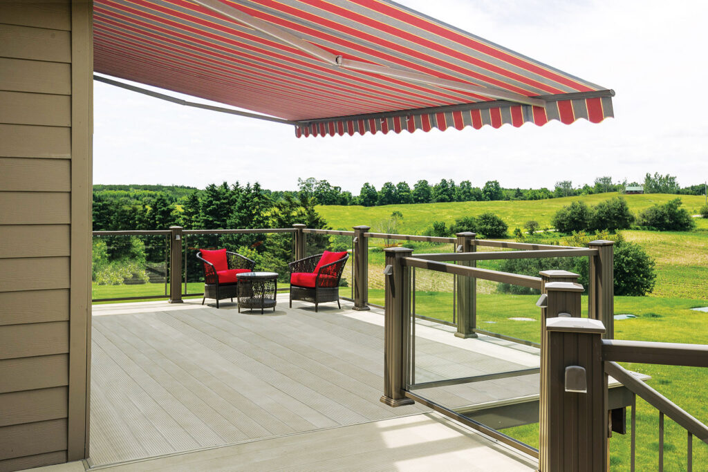 A large Model 1 red and grey striped retractable awning extended over a wide composite deck with glass railings overlooking a green field.