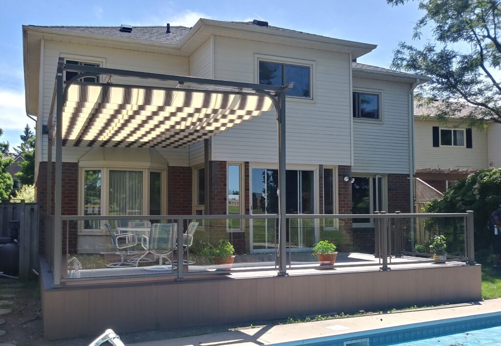 A modern Earthstone-colored aluminum canopy with a black and white striped retractable roof, shading a dining area on a pool deck with glass railings.