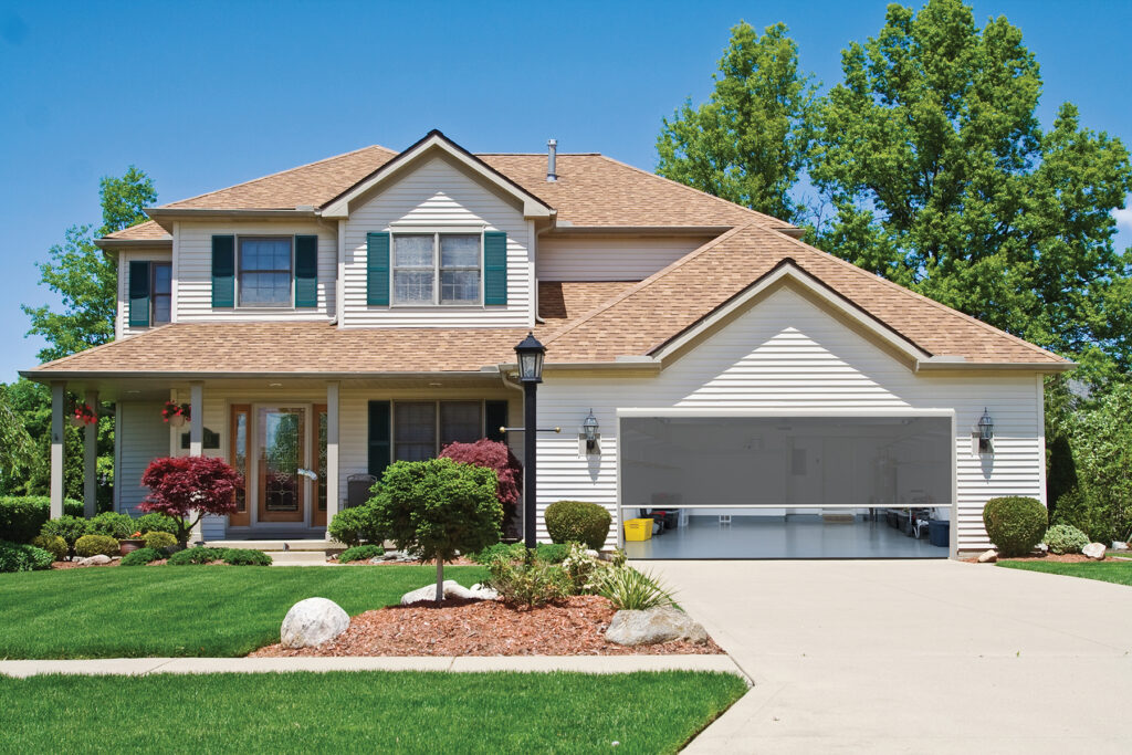 A large motorized solar shade installed over a garage door opening of a suburban home, providing privacy and shade for the interior workspace.
