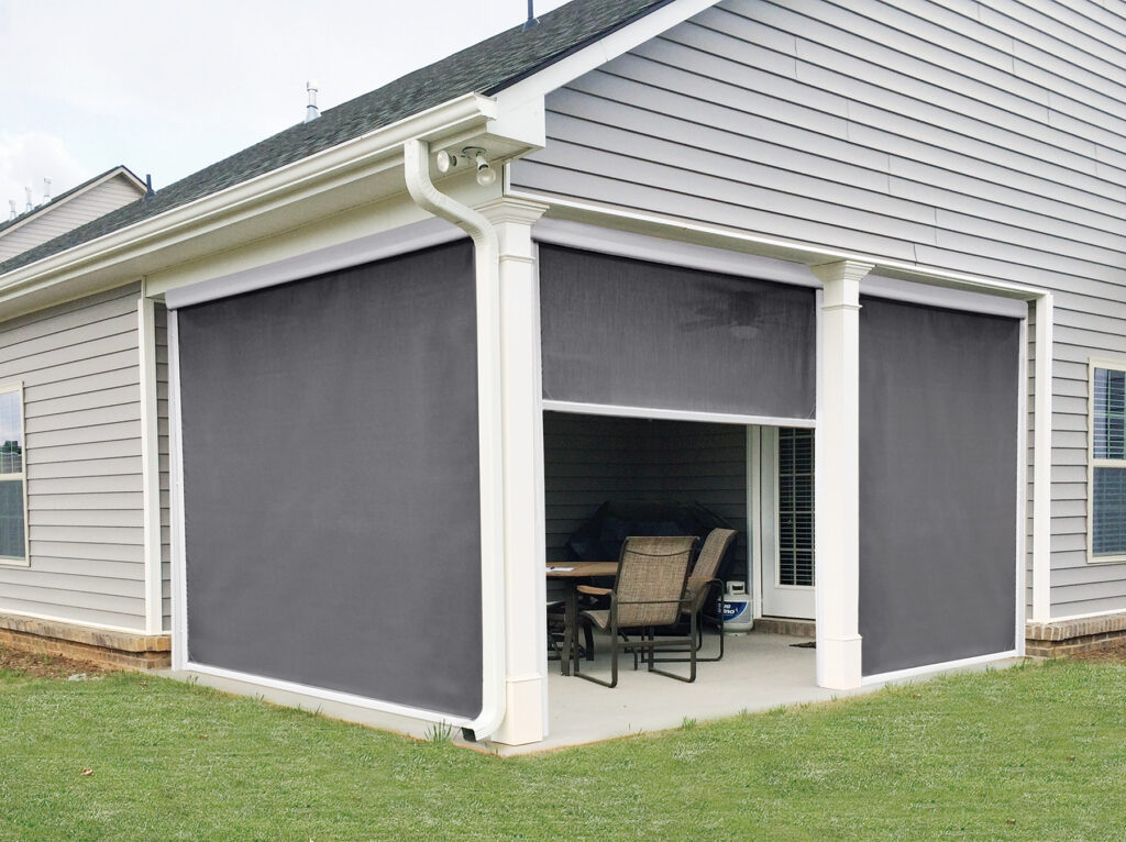 Exterior of a covered patio with grey motorized screens installed between white pillars, showing one screen partially raised to reveal the interior dining area.
