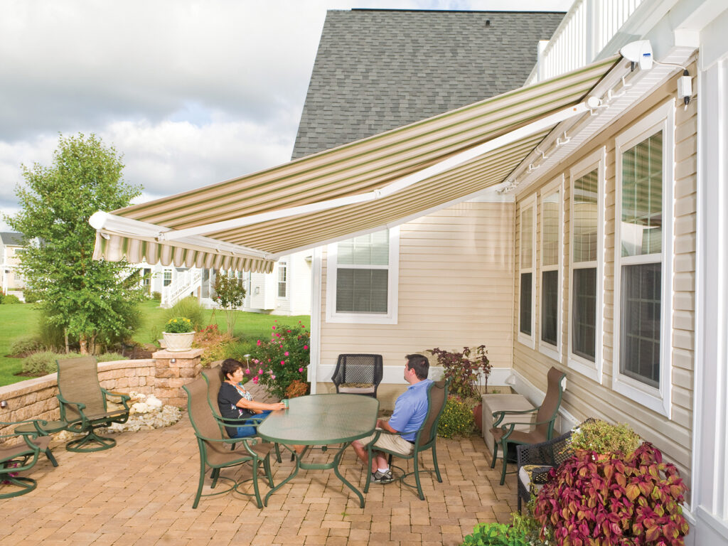 Exterior of a tan home with a striped retractable awning extended over a paver patio where a couple is sitting at an outdoor dining table.
