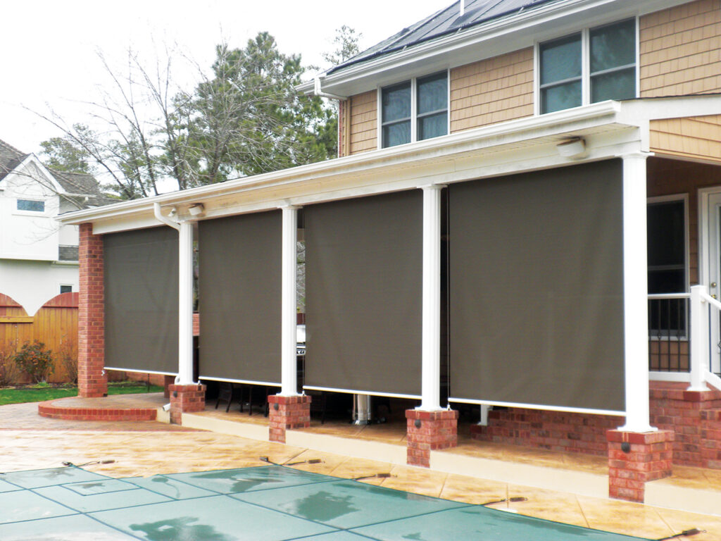 Custom Deck Builder for Homeowners - M2 Decks & Enclosures Exterior of a luxury poolside porch featuring four dark bronze motorized solar shades installed between white columns to block sun glare and heat.
