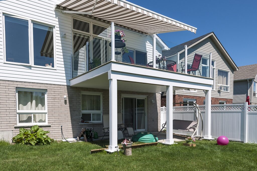 Exterior of an elevated white deck with glass railings and a striped retractable awning extended to provide shade on a bright, sunny day.