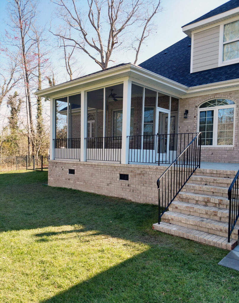 Exterior view of a white aluminum screen room with a brick foundation and black metal railings, showing the open-air design compared to a sunroom.