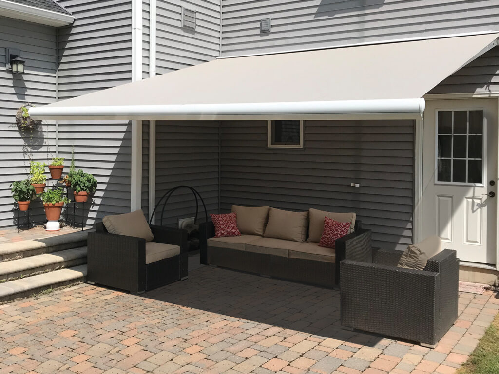 A solid tan motorized awning providing full shade for a grey wicker outdoor sofa set on a brick paver patio next to a house with grey siding.