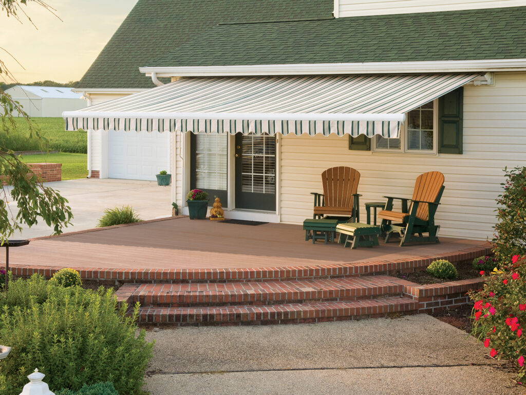 A green and white striped motorized retractable awning extended over a brown composite deck with Adirondack chairs on a sunny afternoon.
