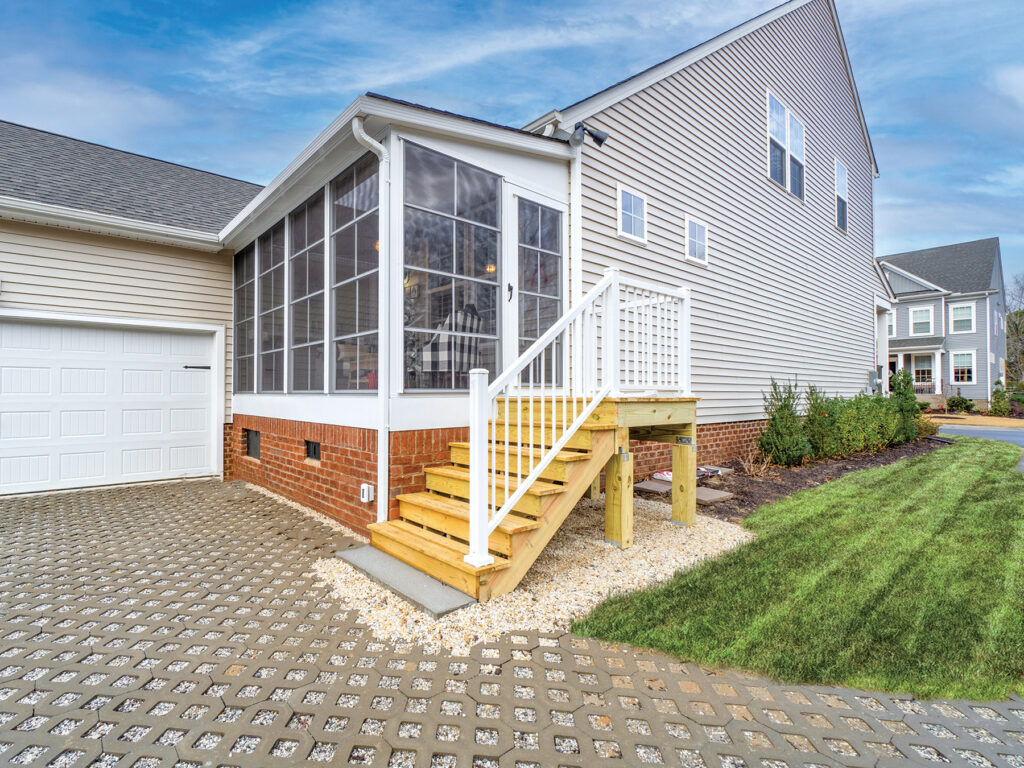 Exterior of a white-framed 3-season room with clear vinyl stacking windows and a brick foundation, attached to a tan siding home.