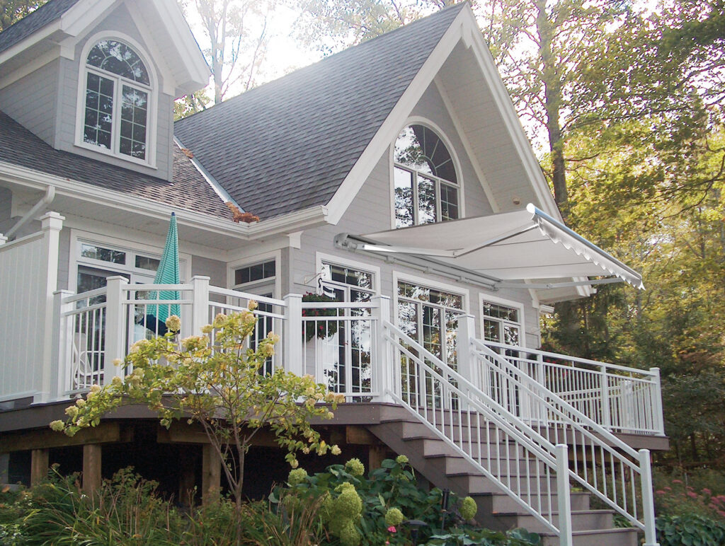 A solid white retractable awning extended over an elevated deck with white railings on a grey gabled home with large arched windows.