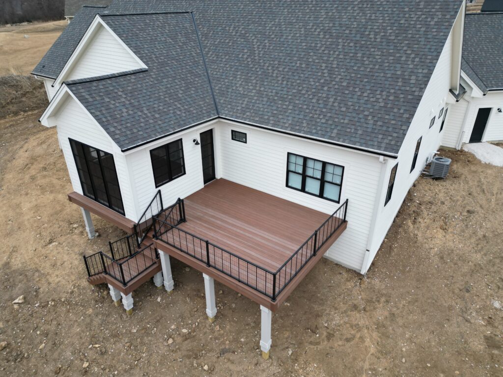 Aerial drone view of a warm brown composite deck with black railings built alongside a white siding house on a muddy construction site.