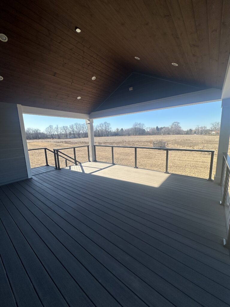 A covered composite deck featuring grey TimberTech decking, black railings, and an integrated wood tongue-and-groove ceiling with recessed lighting overlooking an open field.