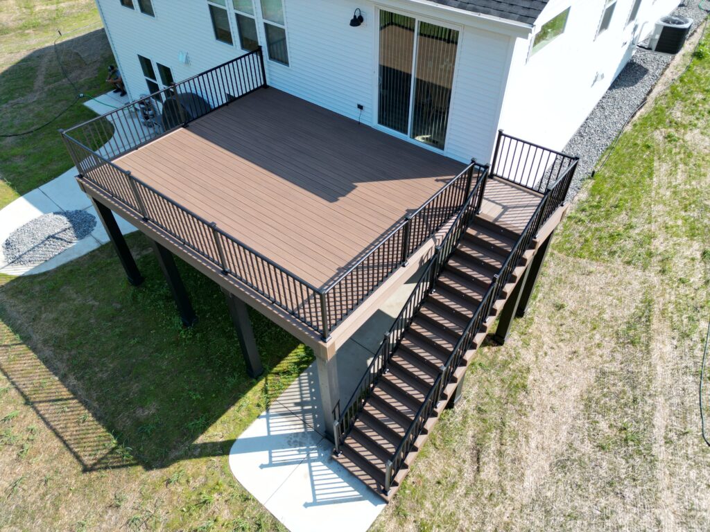 High-angle aerial view of a custom elevated brown TimberTech composite deck with black metal railing and stair system under bright sunlight.